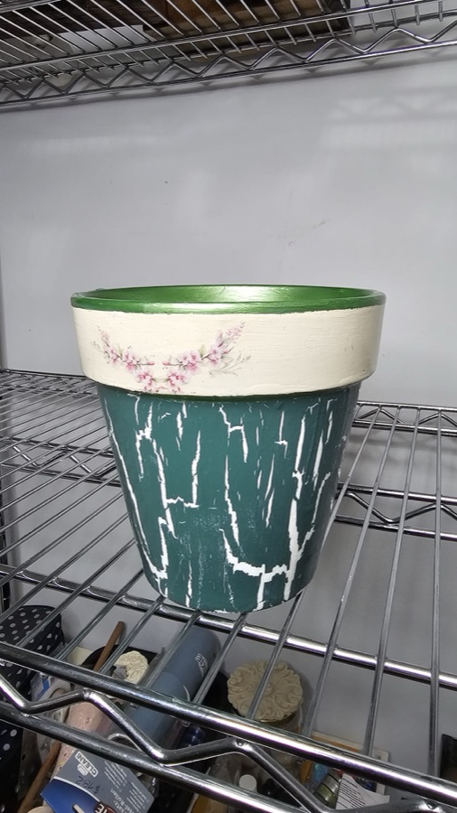 Foto original de una maceta de cerámica con flores pintadas a mano, sacada con el celular sobre una estantería de metal